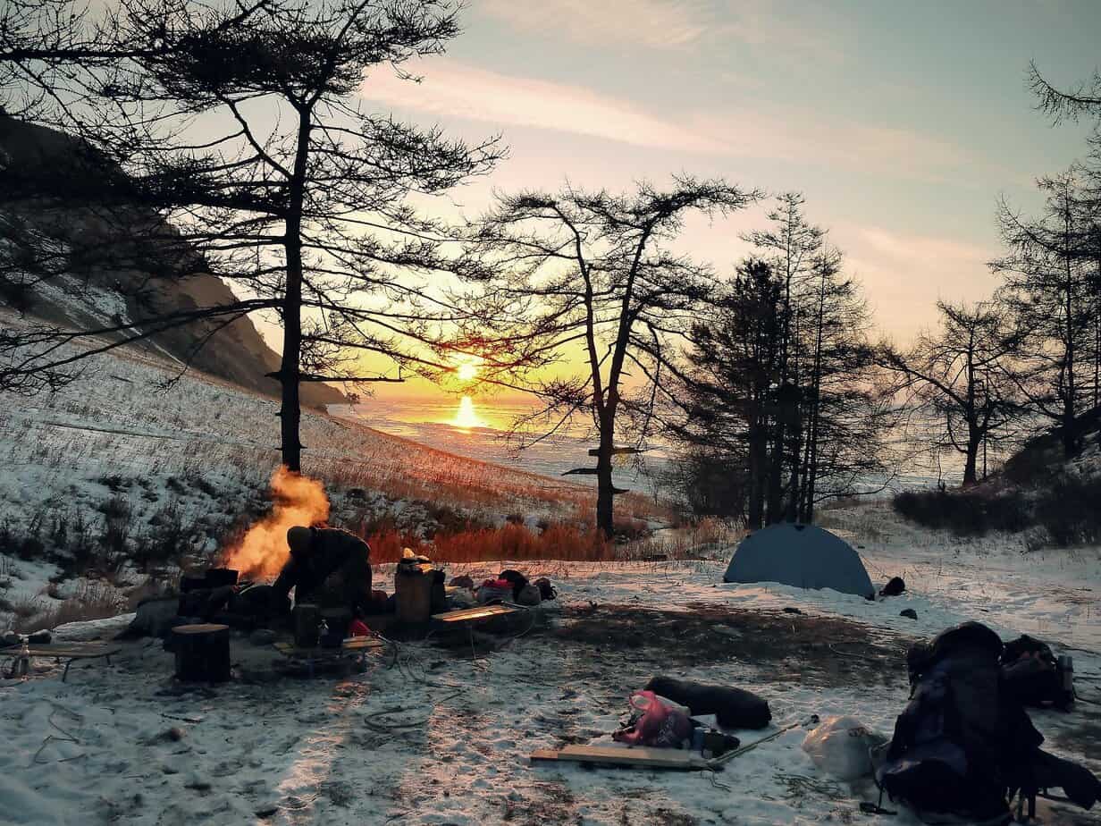 Winter camping dangers shown by a cold-weather campsite with a fire, tent, and heavy snow at sunset.