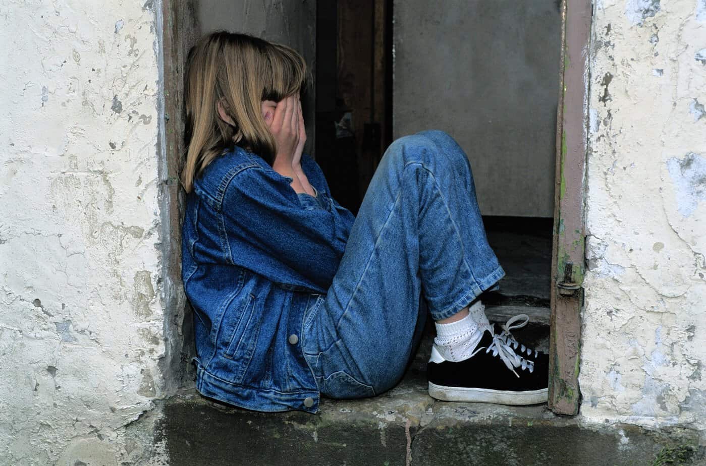 A frightened child sitting alone in a doorway during a crisis, showing the emotional and behavioral stress children may face in emergencies.