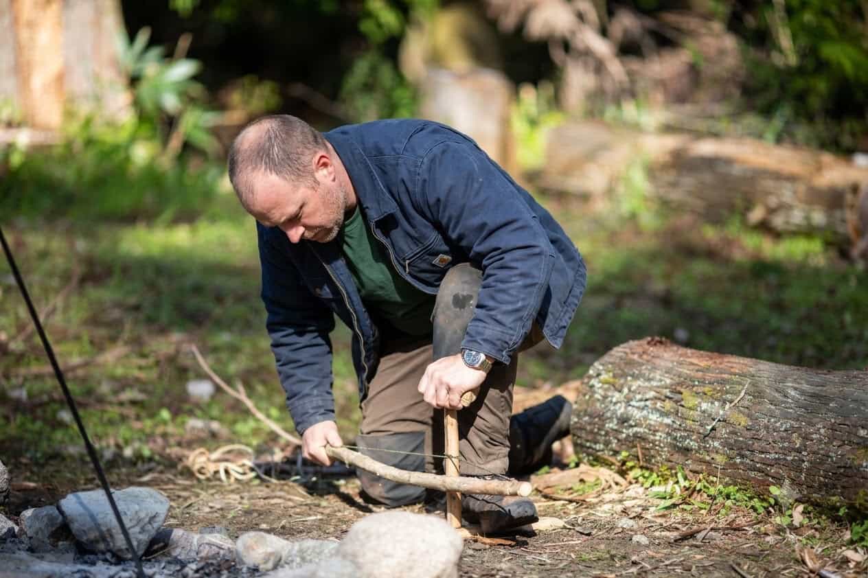 Man kneeling outdoors using a bow drill set to start a friction fire, demonstrating the bow drill deception in real conditions